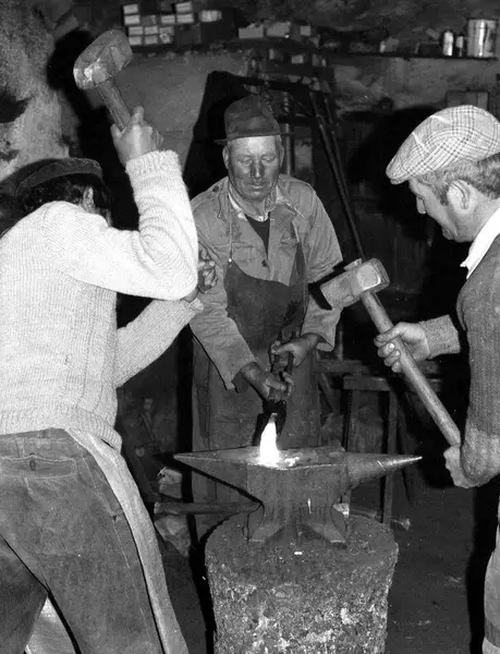 Na frauga: três homens à bigorna, um segura o ferro em brasa e os outros dois malham