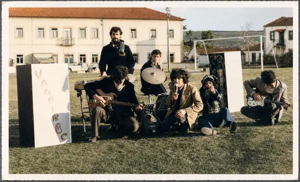Um grupo de rapazes com instrumentos sentados na relva, ao pé de um cartaz