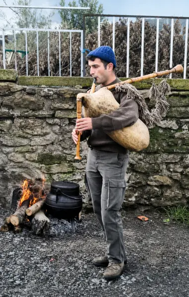 Um gaiteiro de boina a tocar a gaita de foles, ao pé de um lume com um pote